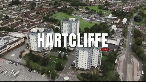 Neil Maggs A bird's eye view of tower block flats and car parks around them. In the distance there are parks, small green fields and houses. The text "Hartcliffe" in white capitals is imposed over the image, along with the text "BS13" in red. 