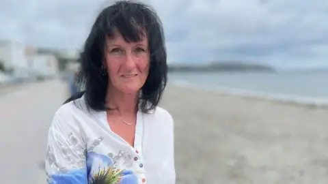 Jackie Hallam, who has shoulder-length black hair and is wearing a white top with a floral design. She is standing on the shore.