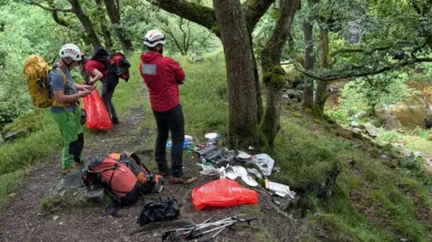 Central Beacons Mountain Rescue Team Two members of the mountain rescue team are wearing rucksacks and are putting litter into bags while another is standing nearby. All three have helmets on. One has the words 'mountain rescue' on the back of his red waterproof jacket.