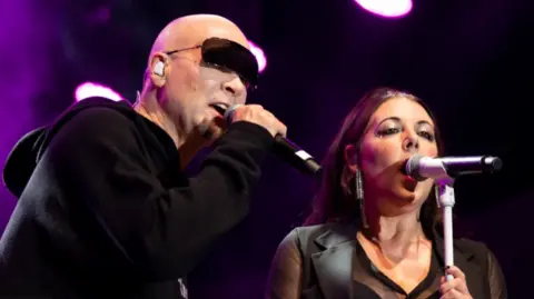 A man, wearing shades and a black hooded top, holds a microphone to his mouth while a woman next to him, wearing a black waistcoat over a sheer top, sings into a microphone on a stand.