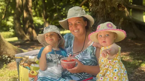 Diamond McGill Diamond McGill sits on a blanket in a wooded area holding a bowl, with her two small children beside her. One child sits on her lap, while the other stands nearby in a sun hat and patterned outfit. Picnic items are placed on the ground next to them.