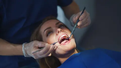 Getty Images a woman has mouth open as her teeth are inspected by a dentis using a scraping tool and a mirror