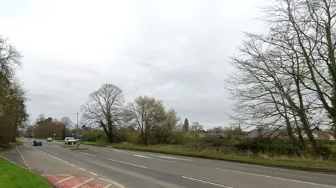 The view across the A49 to the field which can now be developed for housing. Trees line the boundary of the field by the main road.