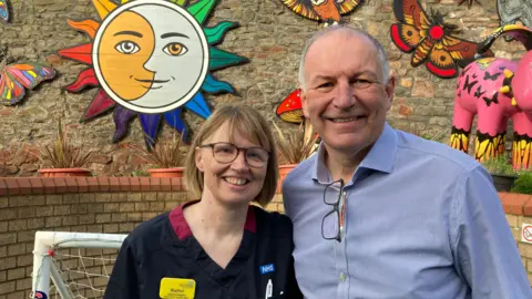 Rachel Hughes and Martin Gargan smile at the camera. Rachel has short brown hair and glasses. She is wearing nurse's scrubs. Martin has short grey hair and is wearing a blue striped shirt with his glasses attached to his shirt. They are standing outside in a kid's play area which is decorated with butterflies, a sun and moon and a Gromit.