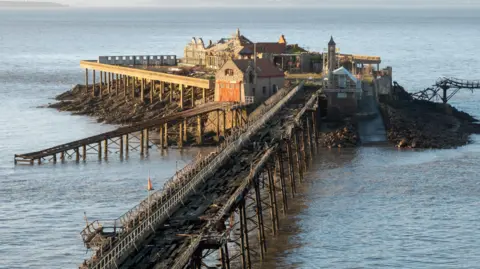 Birnbeck Pier in Weston-super-Mare. The pier is damaged and in disrepair, with a heavily damaged wooden walkway and dilapidated buildings.