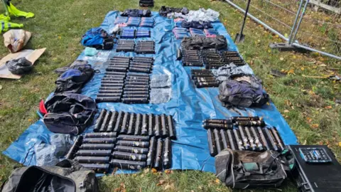 Large pile of black cannisters on a blue tarpaulin in a field with a spade next to it.