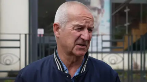Gemma Dillon/BBC A man with a shaven head in a collared shirt and blue jacket speaks to a reporter