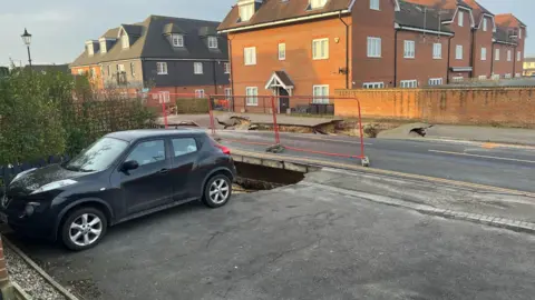 BBC/ADRIAN HARMS A dark car parked in a driveway of a property in Godstone, with the edge of a sinkhole reaching its rear wheels. The original, larger sinkhole is visible on the opposite side of the road, where both one lane of the road and part of the pavement have collapsed. There are a number of houses in the background.