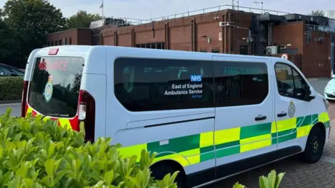 An ambulance outside an ambulance call centre in Bedford. There is a green bush to the left and a red-brick building in the distance.