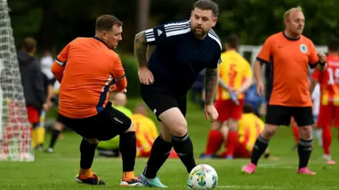 Man v Fat Two men are playing football on a grass pitch, one wearing black kit and the other in orange. There are players from other teams in the background.
