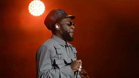 Getty Images Burna Boy wearing a grey outfit, holding a black microphone on stage, with an orange lit background.