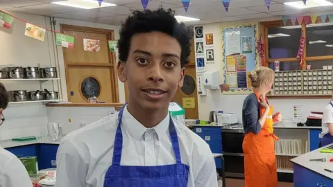 Harrison smiles at the camera in the kitchen classroom. He is wearing a white shirt, buttoned up to the top, and a blue apron. 