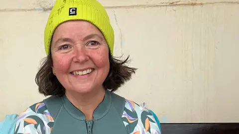 Clare Worden/ BBC Sarah Chant is wearing a green rash vest and a lime green hat. She is ready to go sea swimming and is smiling at the camera. 