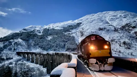 Matt Earnshaw A locomotive crosses a snow-covered viaduct under a clear night sky, with rugged, white-capped mountains rising in the background. 