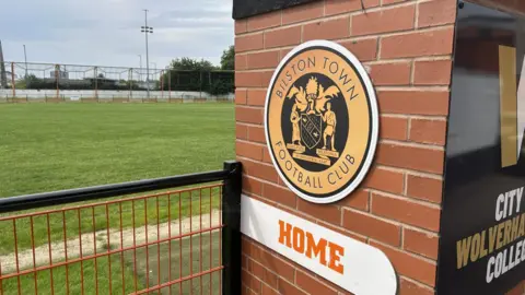 Bilston Town. We can see the pitch, the crest and a "HOME SIGN". It is an overcast day and we can see an orange fence and flood lights in the background.