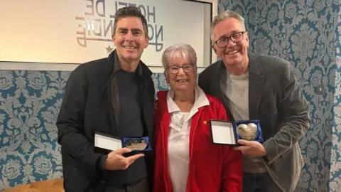Sarah Auckland of Variety Jersey with Greg Wills and James Scott. Wills and Scott are holding silver hearts in a blue case. 