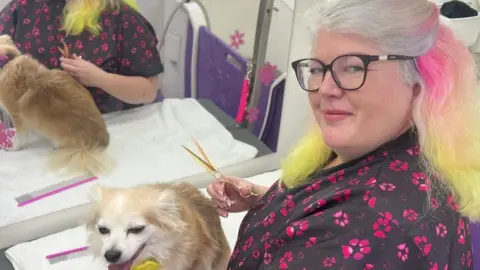 A woman with long hair dyed pink and yellow holds a pair of scissors. A small dog sits on a table in front of her. 