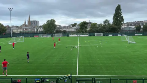 BBC A view of the new 3G football pitch with football players walking across it, footballs in the middle of the pitch and two goals either side. Houses seen afar with grey skies.