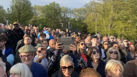 A packed crowd of people standing in an open field looking towards something beyond the camera. One person is holding up a tablet, with another one holding up a phone. Most are wearing sunglasses.