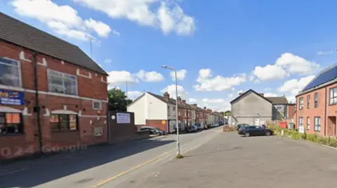 Google A generic photo of a road going from the bottom left to the right side of the image. A two-storey building is on the far left and terraced properties are further down on that side of the road. 