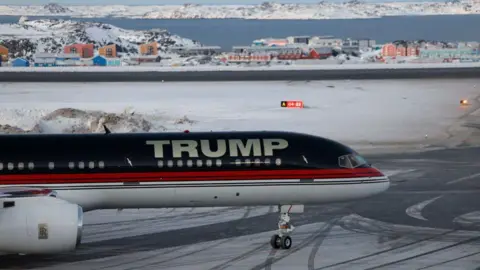 An aircraft carrying President Donald Trump's son, Donald Trump Jr, arrives in Nuuk, Greenland on 7 January 2025. The name "TRUMP" is painted on the plane in large silver letters. Colourful buildings and snow are visible in the background