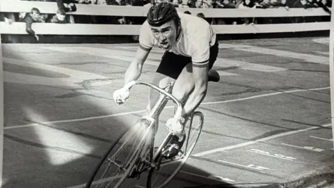 A black and white photo of a man cycling on a track looking strained as crowds cheer him on