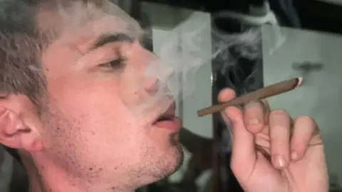 Ross Powell Ross is indoors holding a lit cigar or rolled smoking item between fingers, with visible smoke swirling in the air. The background shows reflective surfaces and muted lighting.