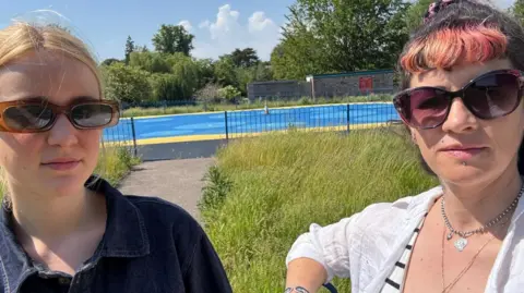 Green Party Livvy Gibbs and Sally Leonard stand in front of a the closed splash park in St Albans posing for a selfie. It is a bright, hot day. 