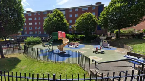 Google A screenshot from Google street view showing a small children's playpark. There are colourful shapes resembling flowers on the ground, and black metal railings surrounding the perimeter. Inside the park there is a small see-saw, a yellow spiral slide, blue metal benches and several climbing frames. The park is surrounded by bushy trees and in the background there is a brown brick block of flats.