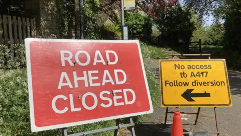 A photo showing a red road closed sign and a yellow traffic sign on a road. In the background there is a bus stop surrounded by trees