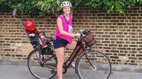 A woman smiling and standing with a bike between her legs. She is wearing a helmet and there is a child on the back with a helmet covering their face.