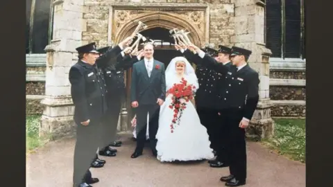 Housden family Mervyn Housden in a grey suit and red buttonhole standing beside Hayley Housden in a long white wedding dress and veil holding a bouquet of red flowers, outside a church with a guard of honour of firefighters on either side in dark uniforms and caps holding hammers aloft