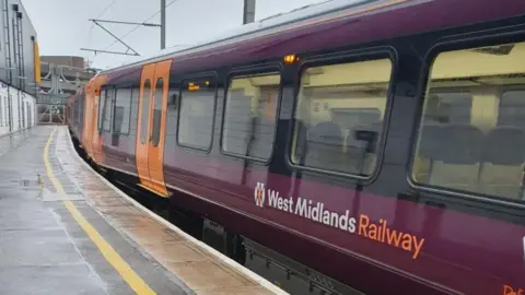 An orange and maroon train at a West Midlands station. There is lettering on the side saying "West Midlands Railway" and the train has multiple windows and several doors. It is next to a platform which has a yellow line down one side.