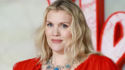 Getty Images Close up of Emerald Fennell on the red carpet. She has blonde, shoulder-length hair and wears a red dress and large necklace featuring blue jewels. 