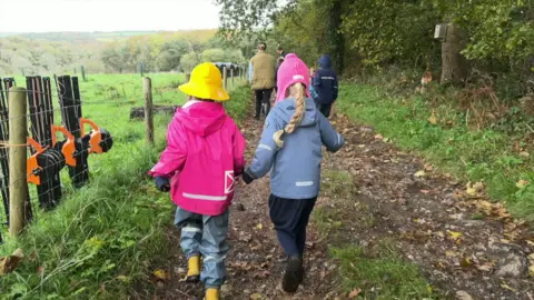 The back of two children walking and holding hands. They are both wearing hats and coats as they walk down the pathway in a field. To the left are wooden posts with metal fencing between them. On the right is grass and trees.