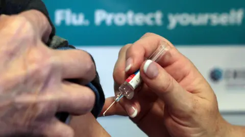 A close-up of someone having an injection. A person is holding the injection in one hand towards an arm. The other person is using their hand to hold up their sleeve.