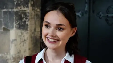 Shaun Whitmore/BBC Jess Clifton is standing in the middle. She has short black hair and is wearing a white shirt and a burgundy dress. She is standing in front of two large black doors.