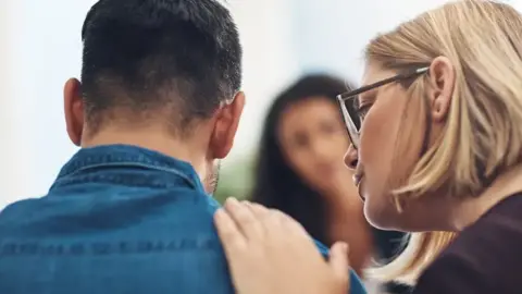 A woman rests her hand on the shoulder of a man with his back to the camera. He is wearing a blue shirt and has dark hair. She has short blonde hair and glasses on.

