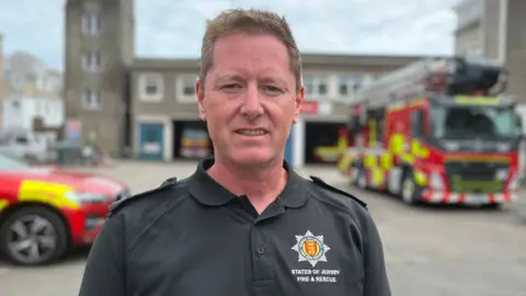 Chris Smith, head of fire prevention at Jersey Fire Service. He is stood in the yard of a fire station. A couple of fire engines are parked up outside. Smith is wearing a black polo shirt with the fire service emblem on it. He has short light brown hair.