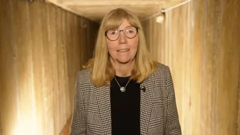 Andrew Marshall/BBC A woman with shoulder-length blonde hair. She is wearing a black shirt with a black and white blazer on top. She is standing in the underground tunnel.