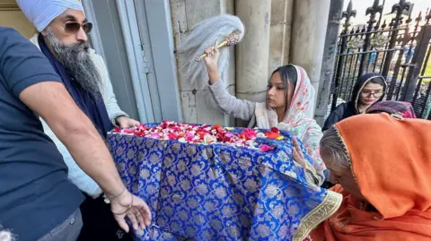 The scripture is inside a box covered in a blue cloth. There are red petals on top.