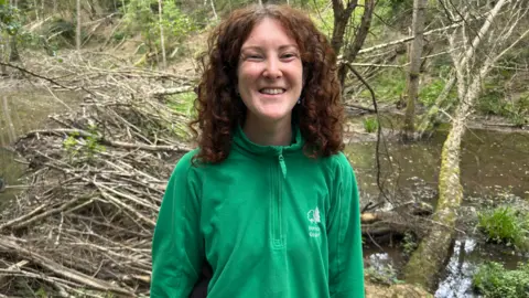 A woman with red curly hair is standing in front of a number of trees in a forest. She is wearing a green jacket with a tree logo and "Forestry England" emblazoned at the front.