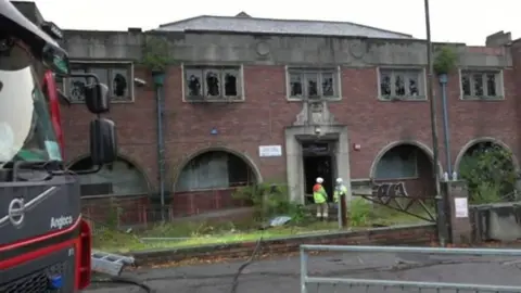 A derelict building with damaged windows and two firefighters outside the property.