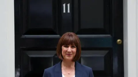 Reuters Rachel Reeves is smiling outside the door of Number 11, which is black with white number 11 on the door above her head. She is wearing red lipstick and she has straight short brown hair and a fringe. She has a small necklace on and is wearing a blue navy blazer. 