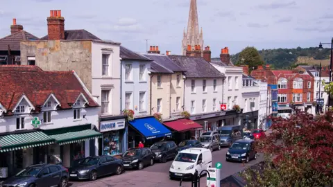 A street scene in Dorking shows cars parked along a road lined with shops, with a church spire rising above buildings in the background.