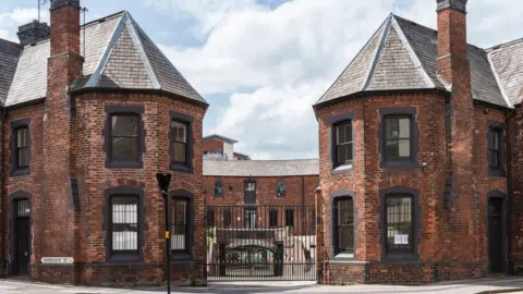 A view of a horse-shoe shaped 18th century brick building with black framed windows and black iron gates at the front 