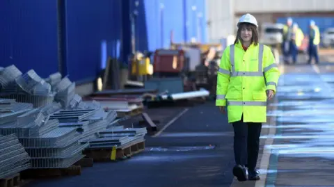 PA Media Rachel Reeves smiles wearing a white hard hat and a yellow fluorescent jacket. She has shoulder length brown hair with a fringe. She walks towards the camera past building parts.