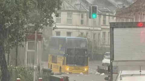 A bus travels through flood water in Plymouth. Heavy rain is pouring down on the yellow and blue double decker buses. Bins have fallen over into the flood water. Other vehicles are attempting to travel on the flooded road. 