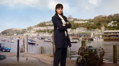 Zahra Ahmadi as the character Det Sgt Esther Williams posing for a press shot with Looe behind her across the water on which yachts are moored. She has long dark hair and is wearing a navy anorak and trousers with a white shirt. She is smiling and has her arms crossed. There are lobster pots at her feet. 