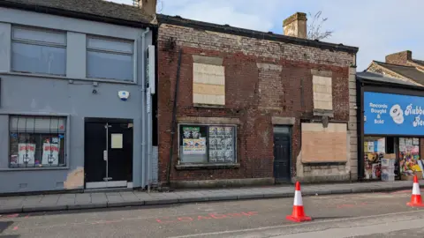 LDRS The front of a building on a city high street. The windows are boarded up and the brickwork is exposed. Two traffic cones are on the road in front of it.